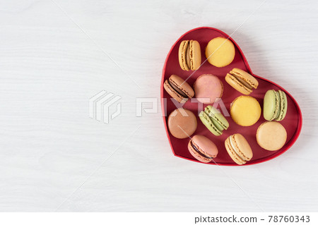 Multi colored french macaroons in a heart shape box top view on white wooden background. Multi colored french macaroons in a heart shape box top view on white wooden background. 78760343