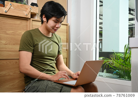 Smiling young man siting in living room and using laptop. Smiling young man siting in living room and using laptop. 78761009