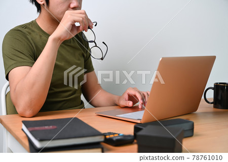Young man holding his glasses and using laptop computer at home. Young man holding his glasses and using laptop computer at home. 78761010