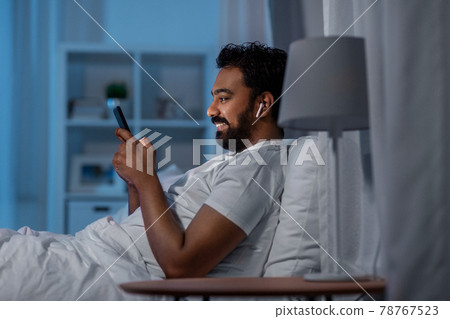 man with smartphone and earphones in bed at night man with smartphone and earphones in bed at night 78767523