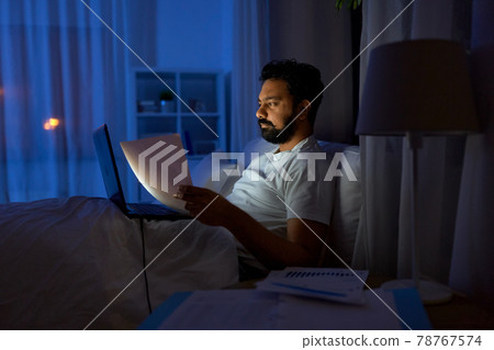 indian man with laptop and papers in bed at night 78767574
