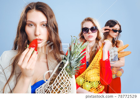 Three young fashionable females housewives in red and blue swimsuits and trench coats on naked body. Shopper strip bags with food in hands. Shopping, purchasing and consumerism. Beautiful long legs. 78772529