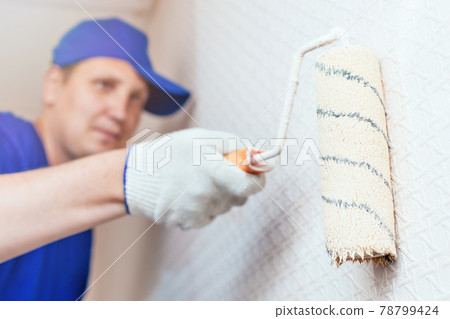 A painter in a cap and gloves paints the wall with a paint roller. Portrait of a young worker in 78799424