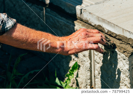 An old bricklayer lays out concrete tiles outside on a summer day. 78799425