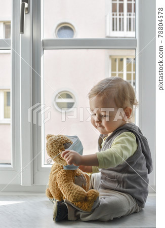 Child in home quarantine playing at the window with his sick teddy bear wearing a medical mask against viruses during coronavirus COVID-2019 and flu outbreak. Children and illness disease concept. Child in home quarantine playing at the window with his sick teddy bear wearing a medical mask against viruses during coronavirus COVID-2019 and flu outbreak. Children and illness disease concept. 78804578