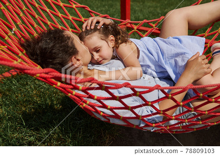 Young mother gently hugs her little daughter lying together on a hammock, enjoying the time in a place on a nature on a warm sunny summer day 78809103