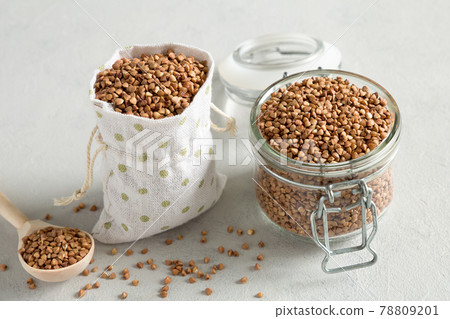 Raw buckwheat grain in a glass jar and cereals in a canvas bag on a gray concrete background. Wooden spoon with grits. Methods of preservation of grains. Healthy, organic, dietary food. Raw buckwheat grain in a glass jar and cereals in a canvas bag on a gray concrete background. Wooden spoon with grits. Methods of preservation of grains. Healthy, organic, dietary food. 78809201
