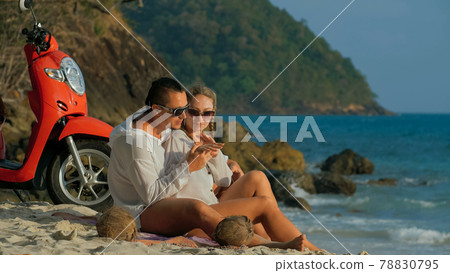 Scooter road trip. Lovely couple on red motorbike in white clothes on sand beach. People walking near the tropical palm trees, sea. Motorcycle rent. Scooter road trip. Lovely couple on red motorbike in white clothes on sand beach. People walking near the tropical palm trees, sea. Motorcycle rent. 78830795