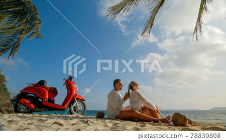Scooter road trip. Lovely couple on red motorbike in white clothes on sand beach. People walking near the tropical palm trees, sea. Motorcycle rent. 78830808