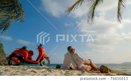 Scooter road trip. Lovely couple on red motorbike in white clothes on sand beach. People walking near the tropical palm trees, sea. Motorcycle rent. 78830813