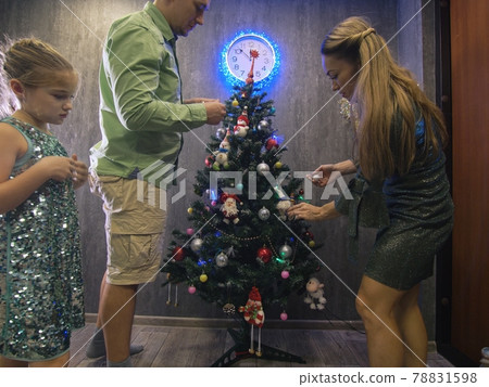 The family fast and fanny decorates the Christmas tree. Side view. Time on the clock is 23.45 on wall. The family fast and fanny decorates the Christmas tree. Side view. Time on the clock is 23.45 on wall. 78831598