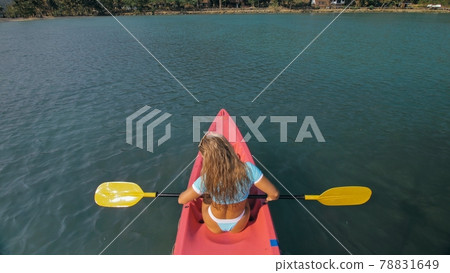 Seductive blonde woman in blue swimsuit sails on pink plastic kayak along azure sea at exotic resort backside upper above view. 78831649