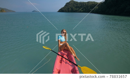Pretty sportive blonde lady in stylish swimsuit sails on pink plastic canoe along azure ocean bay at exotic resort. 78831657
