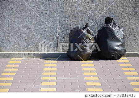 Two black garbage bags on tiled street floor at concrete fence in city Two black garbage bags on tiled street floor at concrete fence in city 78832435