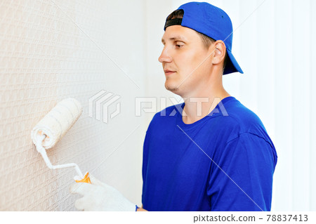 A painter in a cap and gloves paints the wall with a paint roller. Portrait of a young worker in 78837413