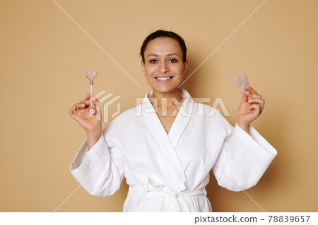 Smiling middle aged woman in white waffle bathrobe posing with gua sha and jade roller massager in her hands isolated over beige background with copy space Smiling middle aged woman in white waffle bathrobe posing with gua sha and jade roller massager in her hands isolated over beige background with copy space 78839657