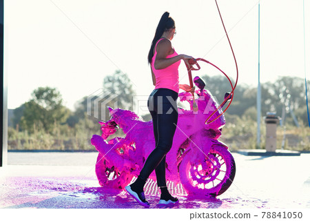 Stunning girl washes motorcycle in car wash service with high pressure water. Stunning girl washes motorcycle in car wash service with high pressure water. 78841050