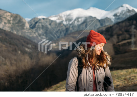 A portrait of a smiling traveler at the snowy mountains background. Happy woman in red cap, warming up and enjoying the sunlight while hiking in the mountains. 78854417