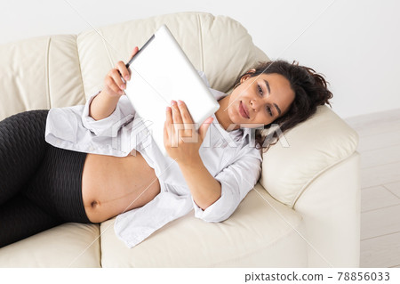 Pregnant woman holding tablet sitting on a carpet near a couch in the living room at home Pregnant woman holding tablet sitting on a carpet near a couch in the living room at home 78856033