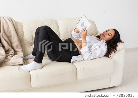 Pregnant woman holding tablet sitting on a carpet near a couch in the living room at home Pregnant woman holding tablet sitting on a carpet near a couch in the living room at home 78856035