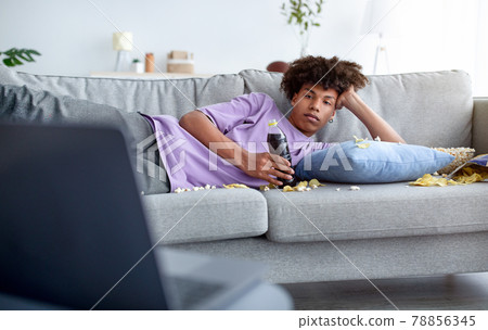 Bored teen guy lying on sofa with scattered snacks, watching movie on laptop at home 78856345