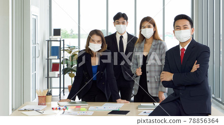 Asian businessman in a suit leaning against the table with a confident expression. The team is preparing for the meeting in the background. Everyone wears a mask. Concept of leadership. Asian businessman in a suit leaning against the table with a confident expression. The team is preparing for the meeting in the background. Everyone wears a mask. Concept of leadership. 78856364
