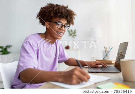 Home schooling concept. Smart black teen guy using laptop at table, writing in notebook during online lesson indoors Home schooling concept. Smart black teen guy using laptop at table, writing in notebook during online lesson indoors 78856419