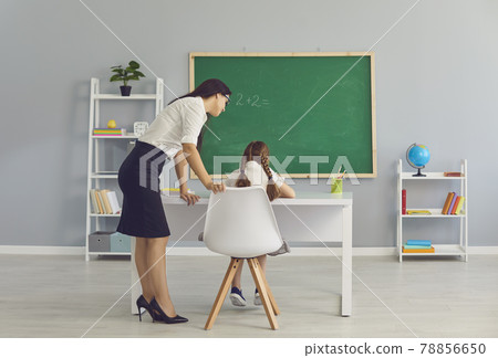 Teacher standing and checking girl pupil sitting and doing homework in school 78856650