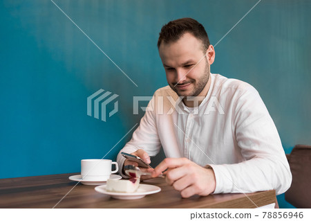 Young attractive businessman in a white shirt sits at a table in the phone and smiles, drinks coffee and eats cake on a blue background 78856946