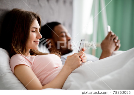Gadget addiction concept. Young multiethnic family using smartphones while lying in bed, copy space 78863095