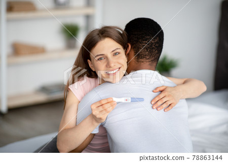 Married multiracial couple happy with positive result of pregnancy test, hugging each other at home 78863144