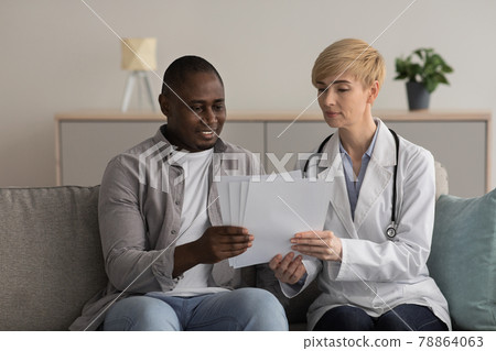 Serious woman doctor in white coat talks about treatment to happy 30s african american guy disabled sitting on couch 78864063