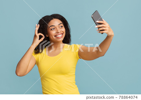 Selfie fun. Happy african american woman taking self-portrait on smartphone and showing ok gesture, blue background 78864074
