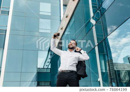Business win. Handsome businessman shaking fists celebrating success at work, standing against office building 78864199