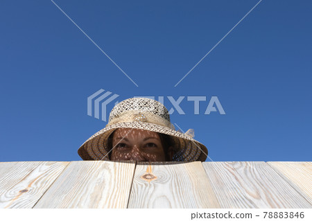 Happy woman sticking out from behind wooden fence or table against blue sky. 78883846