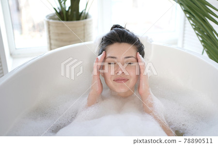 Attractive multi ethnic girl relaxing in bathtub touching healthy soft face skin after daily treatment. Young mixed race asian woman doing massage satisfied with facial procedures skincare concept. 78890511