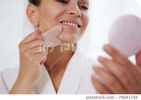 Focus on lips of a beautiful woman looking at a mirror during massaging her face with gua-sha stone massager 78891298