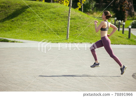 Young female runner, athlete is jogging in the city street in spring sunshine. Beautiful caucasian woman training, listening to music. Concept of sport, healthy lifestyle, movement, activity. Young female runner, athlete is jogging in the city street in spring sunshine. Beautiful caucasian woman training, listening to music. Concept of sport, healthy lifestyle, movement, activity. 78891966