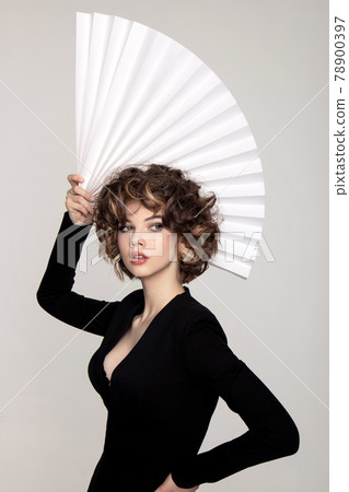 Beautiful european girl with curls with a fan in black dress on a white background 78900397