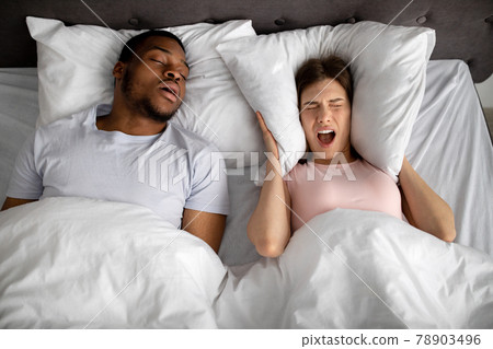 Top view of snoring black husband and his stressed wife screaming and covering ears with pillow in bed at home 78903496