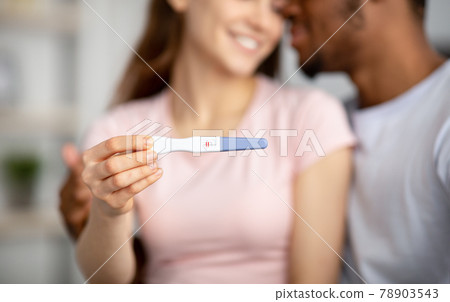 Closeup of happy young multiracial couple showing positive pregnancy test at camera, happy to have baby, selective focus Closeup of happy young multiracial couple showing positive pregnancy test at camera, happy to have baby, selective focus 78903543