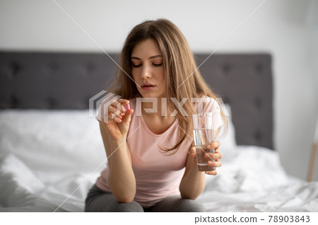 Millennial lady with glass of water taking pill on bed, treating her illness at home Millennial lady with glass of water taking pill on bed, treating her illness at home 78903843