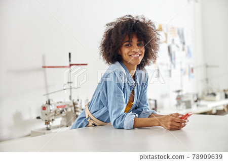 African-American seamstress with smartphone rests leaning onto cutting table in workshop 78909639