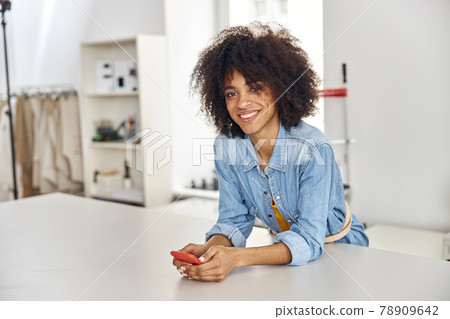 African-American seamstress holds cellphone leaning onto large cutting table in workshop 78909642