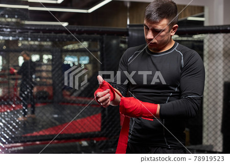 Confident Caucasian Male Kickboxer Fighter Preparing For Fight, Wrapping Hand In Red Bandage Confident Caucasian Male Kickboxer Fighter Preparing For Fight, Wrapping Hand In Red Bandage 78919523