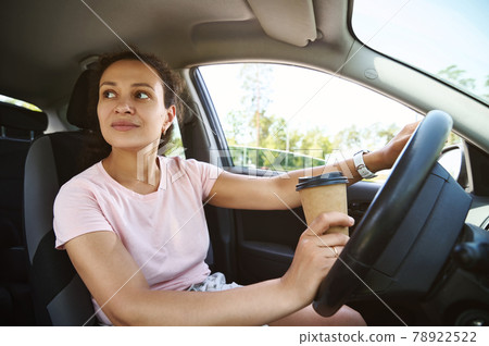 Young unfastened woman driver looking to the side while driving a car and drinking coffee from a takeaway cup 78922522
