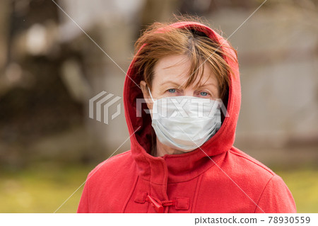 An elderly woman with brown hair and blue eyes in a red coat and hood in a protective safe medical mask, close-up portrait An elderly woman with brown hair and blue eyes in a red coat and hood in a protective safe medical mask, close-up portrait 78930559