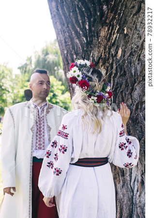 Young people are posing in traditional folk costumes. Cossacks man and woman in wreaths and embroidered shirts. Reconstruction of vintage traditions and customs. Guy and girl kiss 78932217