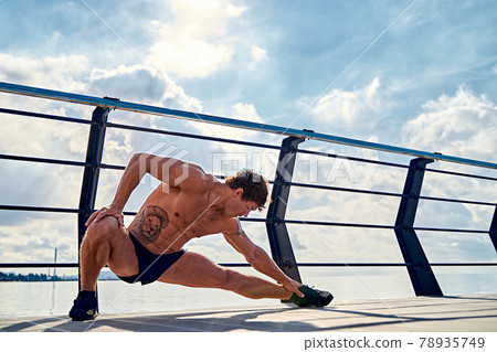 Young muscular tattooed man doing exercises, stretching early in the morning on pier by the sea 78935749