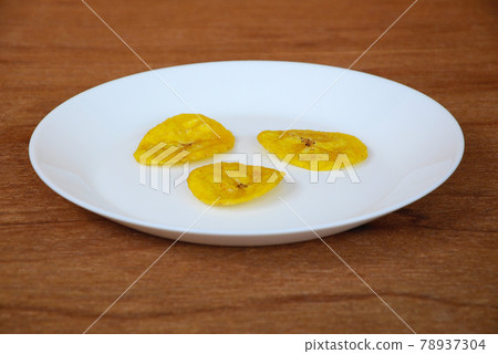 Dried and fried banana slices place on white plate isolated on wood texture background. This is local Malaysian traditional junk food call Kerepek Pisang.    Dried and fried banana slices place on white plate isolated on wood texture background. This is local Malaysian traditional junk food call Kerepek Pisang.    78937304